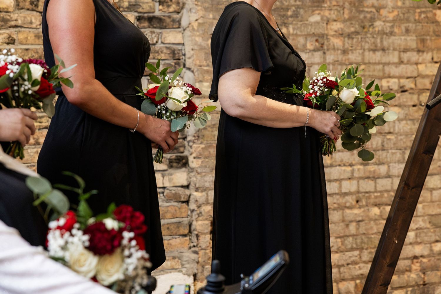 Bridesmaids in black dresses holding red and white floral bouquets at a wedding ceremony near a brick wall.