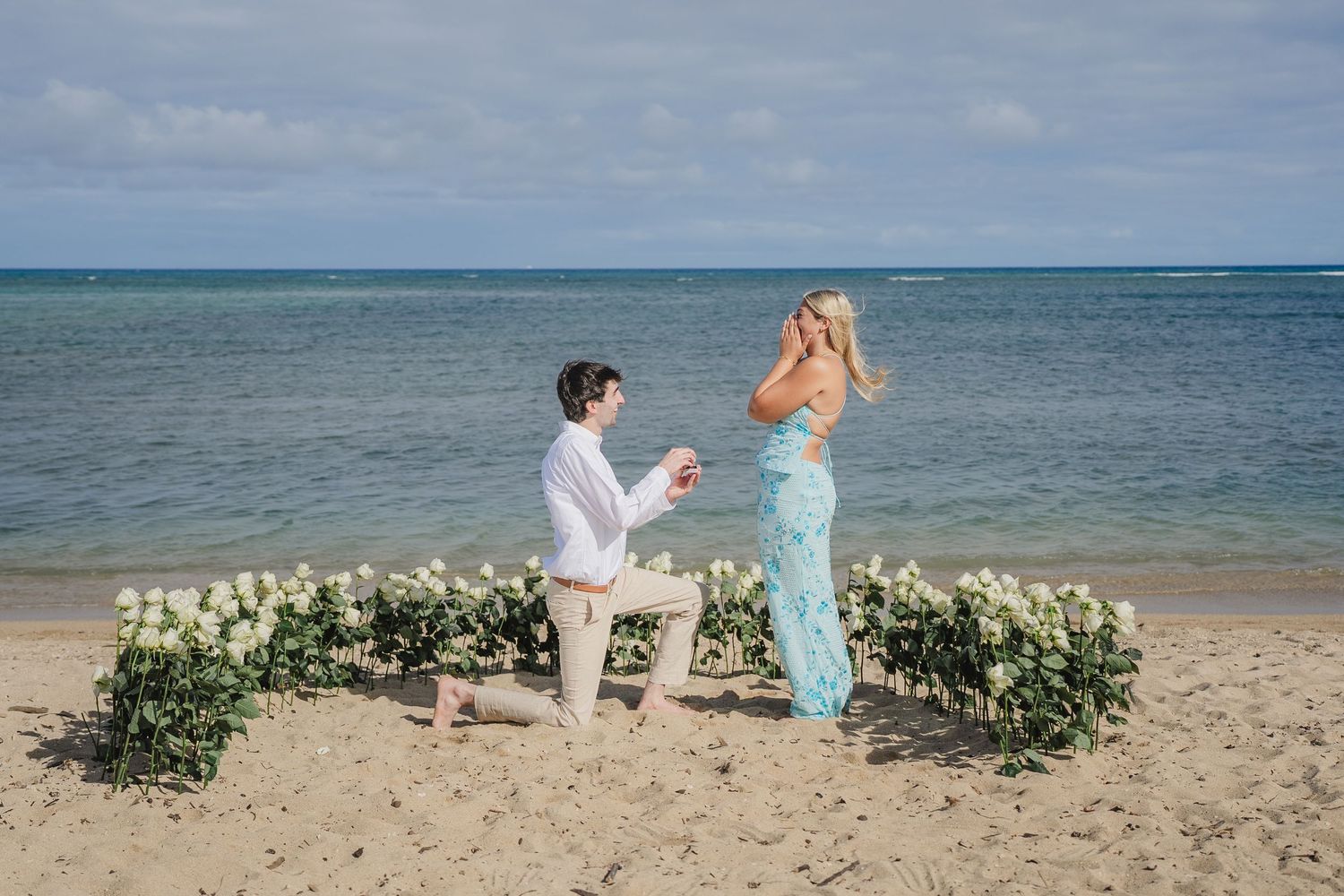 Man proposes to woman on sandy beach with floral arrangement, ocean backdrop, romantic engagement moment in Hawaii.