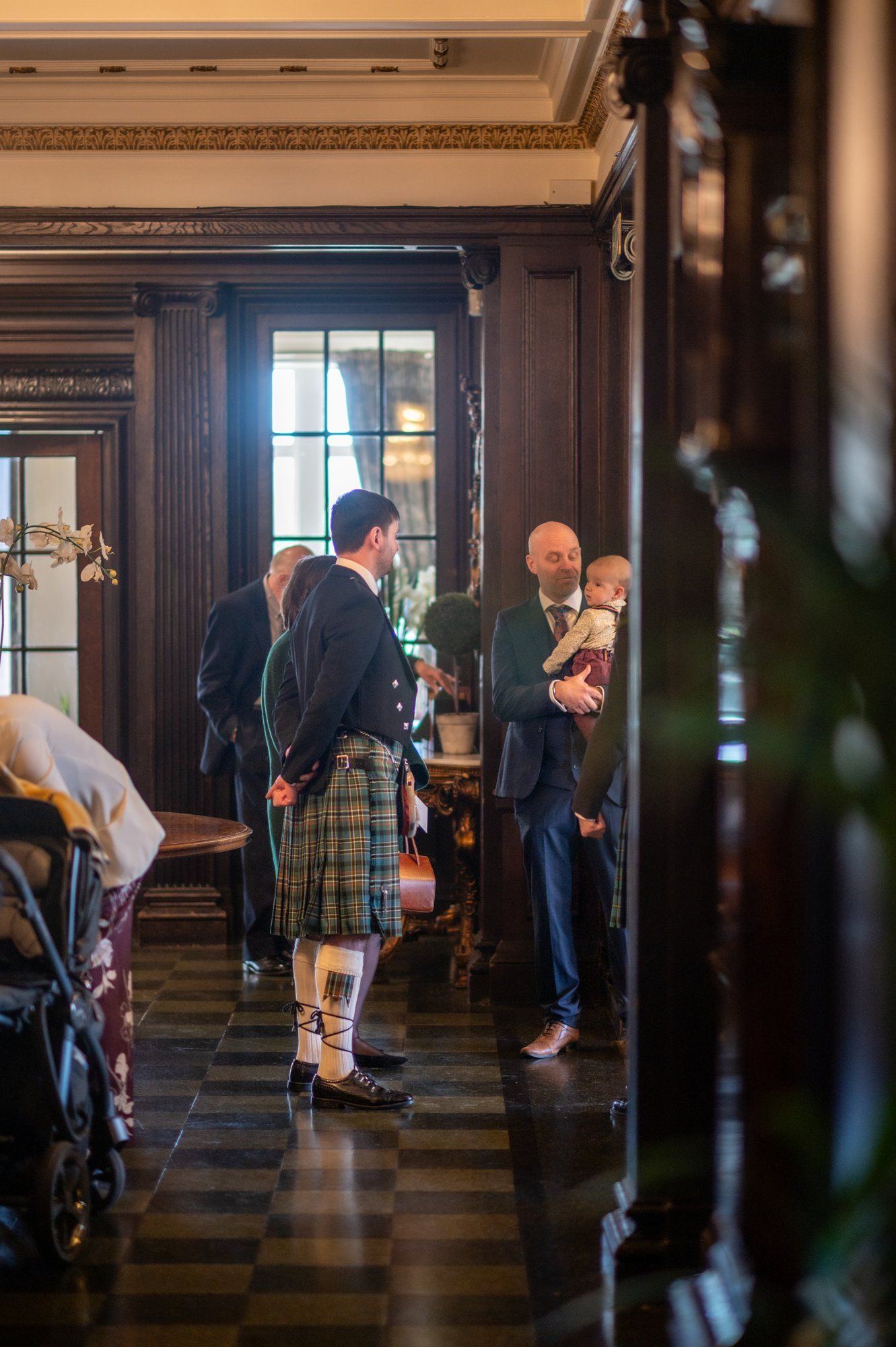 Man in traditional Scottish kilt converses with a guest holding a baby in an elegant wood-paneled hall.