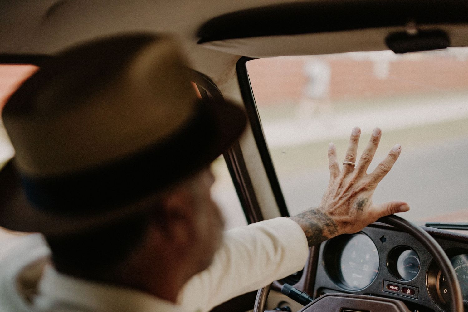 Tattooed man wearing a fedora hat driving a vintage car with hand resting on the steering wheel.