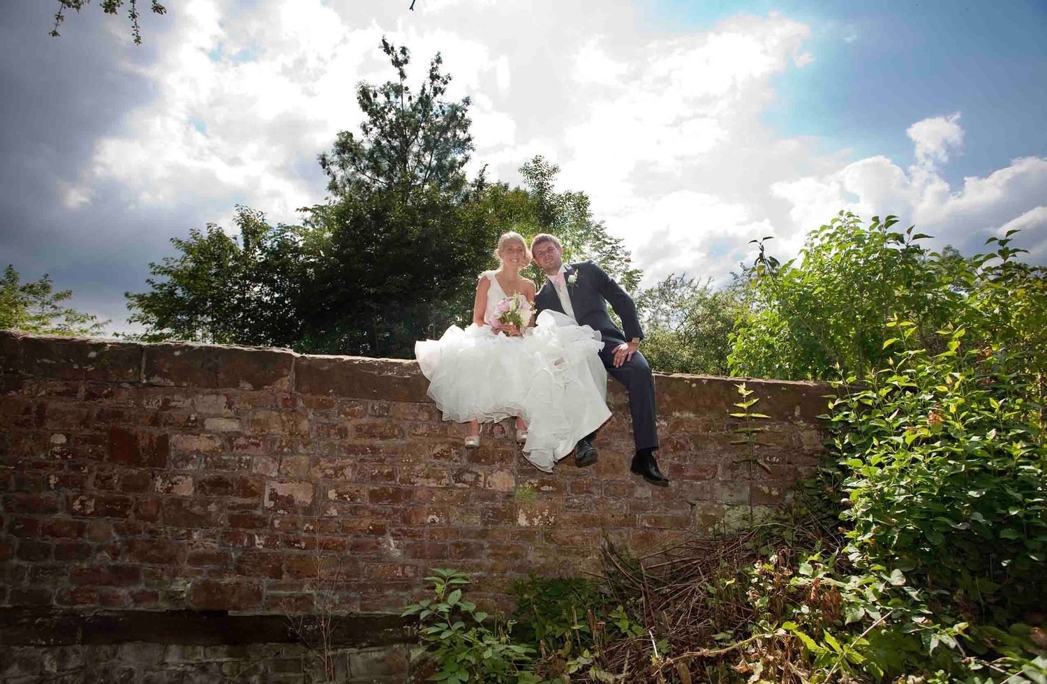 Bride and groom sitting together on a red brick garden wall at Heather Glen, surrounded by greenery beneath a cloud-filled sky.