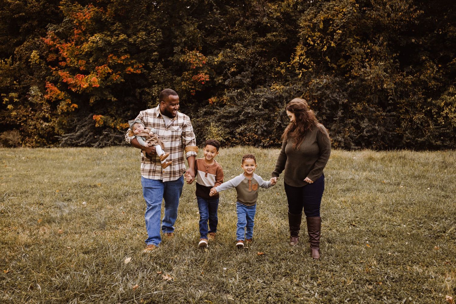 Family swinging child between them in autumn field near rustic cabin, laughing together outdoors.