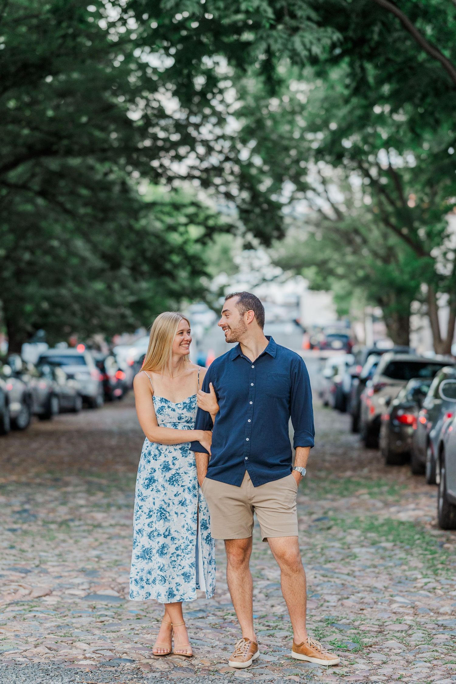 Couple walking on cobblestone street lined with trees and parked cars, woman in floral dress, man in navy shirt.