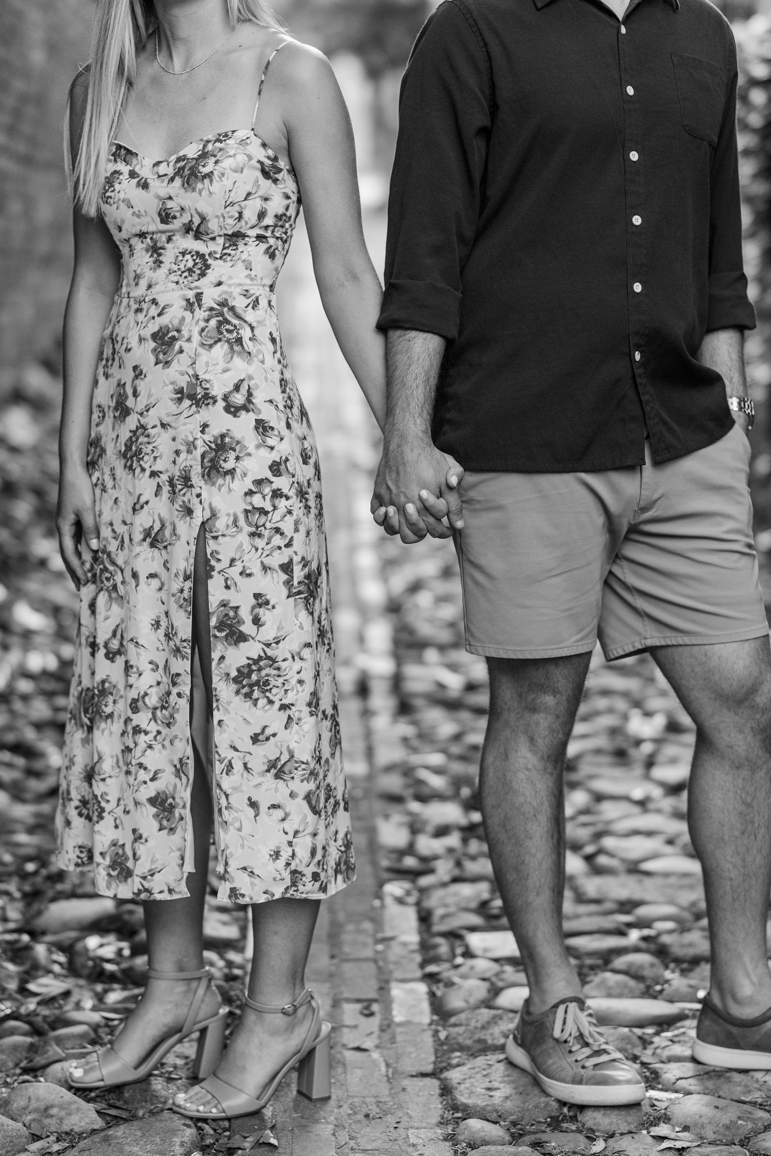 Black and white photo of a couple holding hands, woman in floral midi dress and heels, man in shorts on cobblestone path.