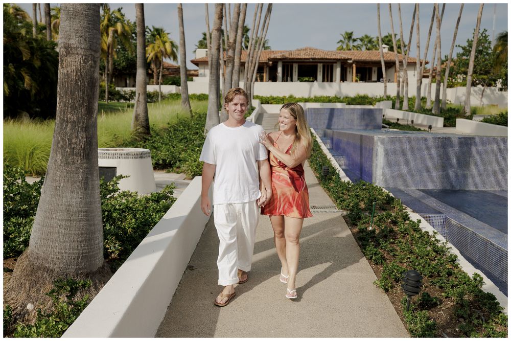 Punta Mita Proposal Photography featuring a couple walking hand in hand along a palm-lined pathway at a tropical resort.