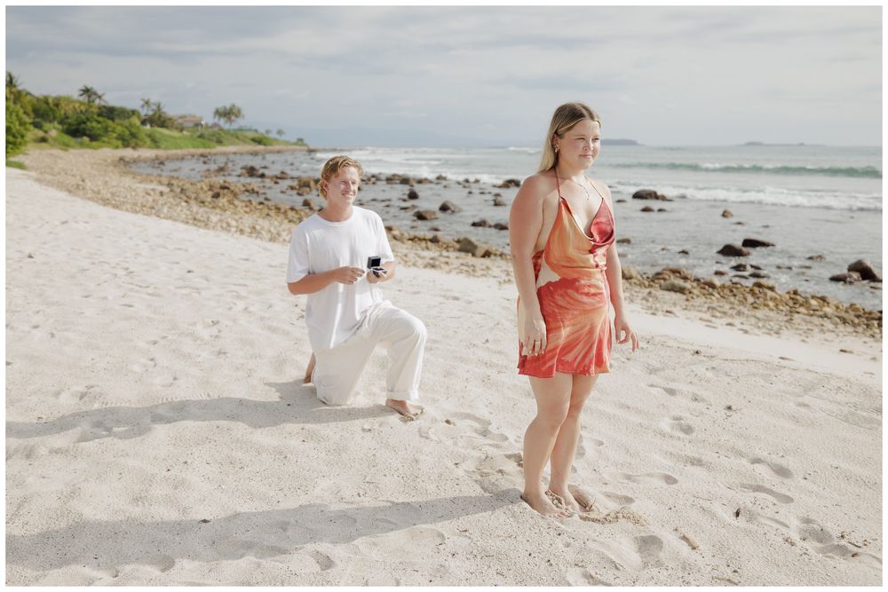Punta Mita Proposal Photography featuring a Groom kneeling on the white sand as he proposes to his bride-to-be, with ocean waves in the back