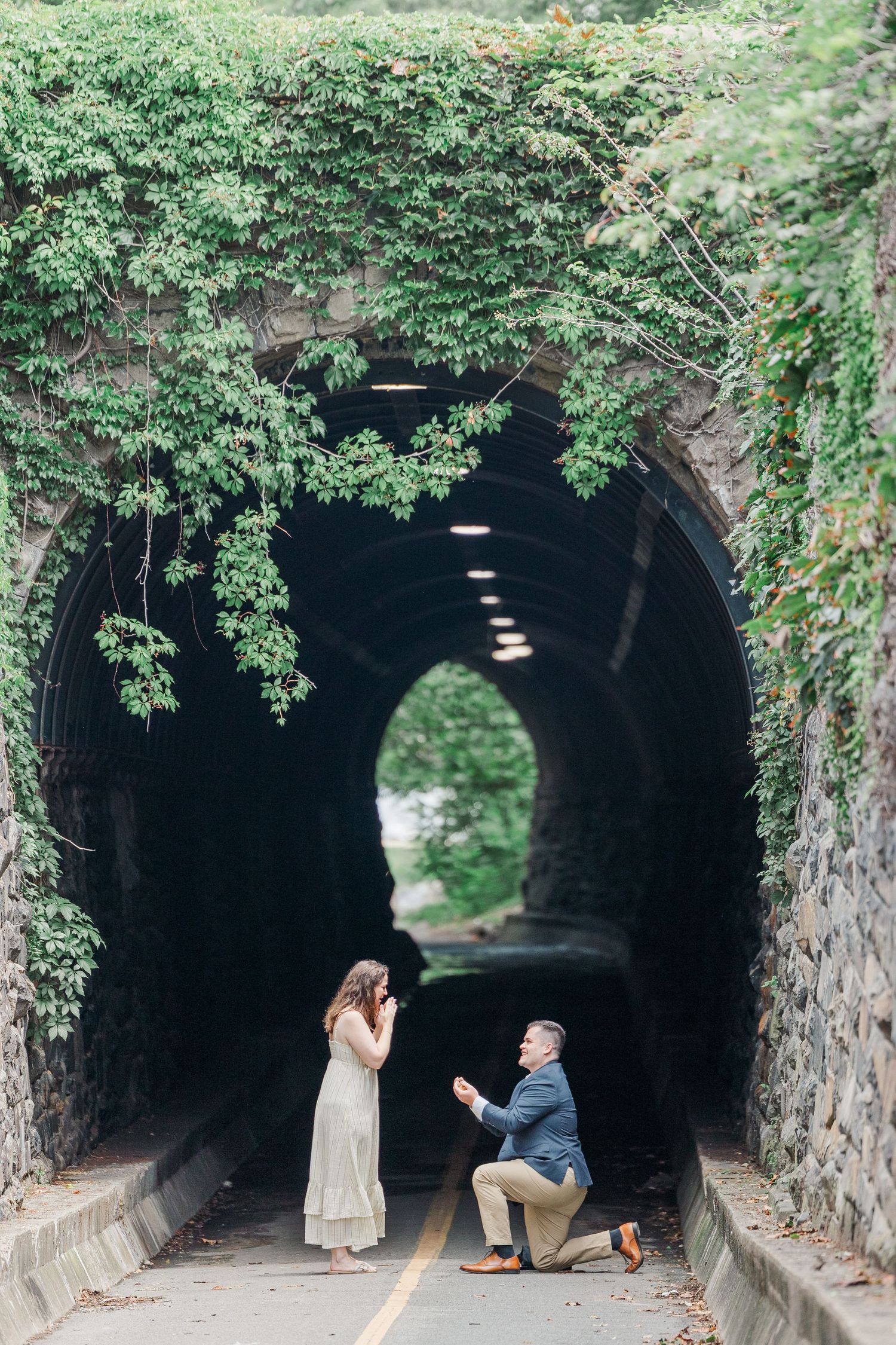 Man proposes on one knee to woman at ivy-covered stone tunnel entrance, romantic outdoor engagement moment.