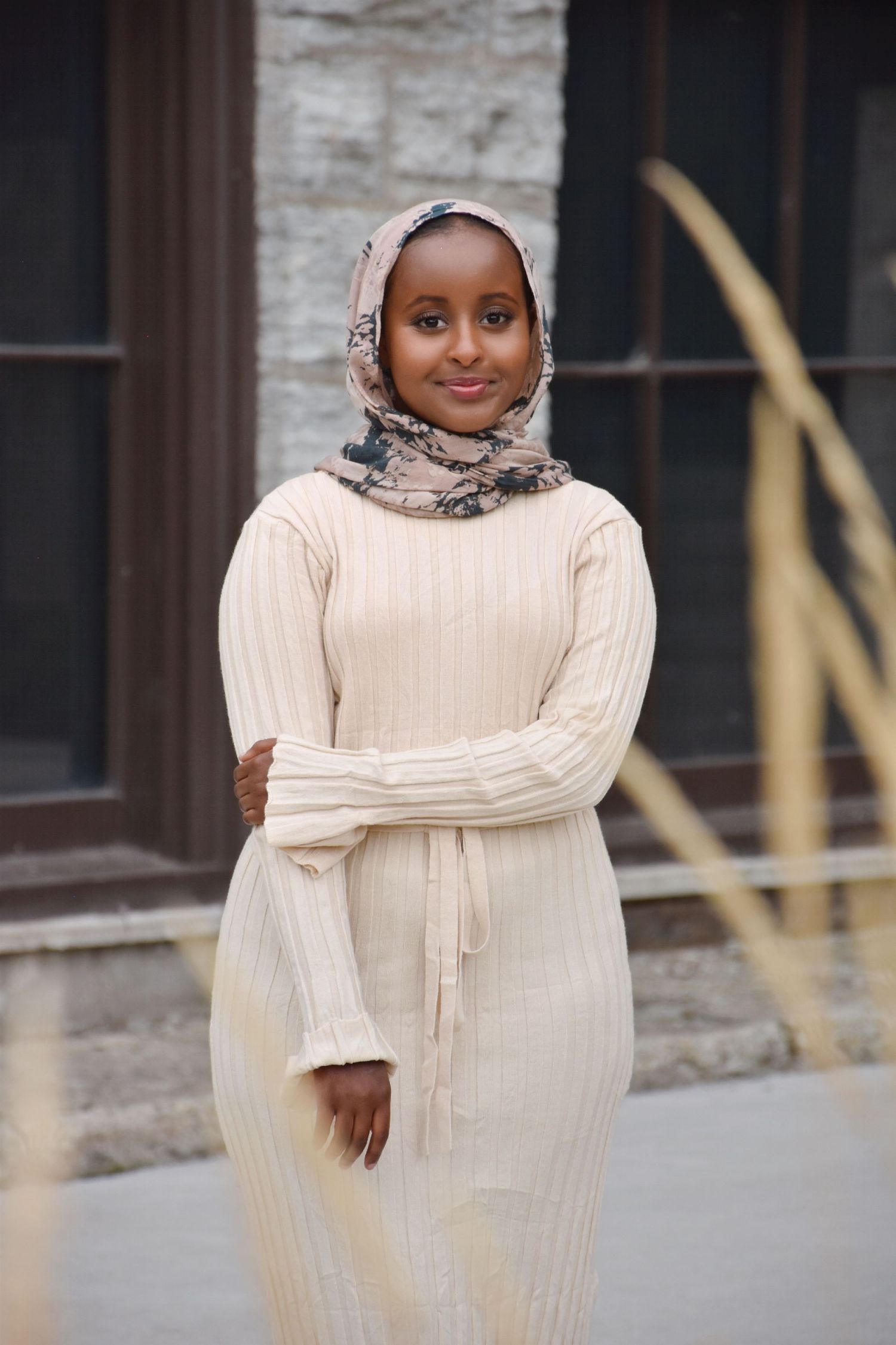 Young woman in cream ribbed dress and floral hijab smiling outdoors near a stone building.