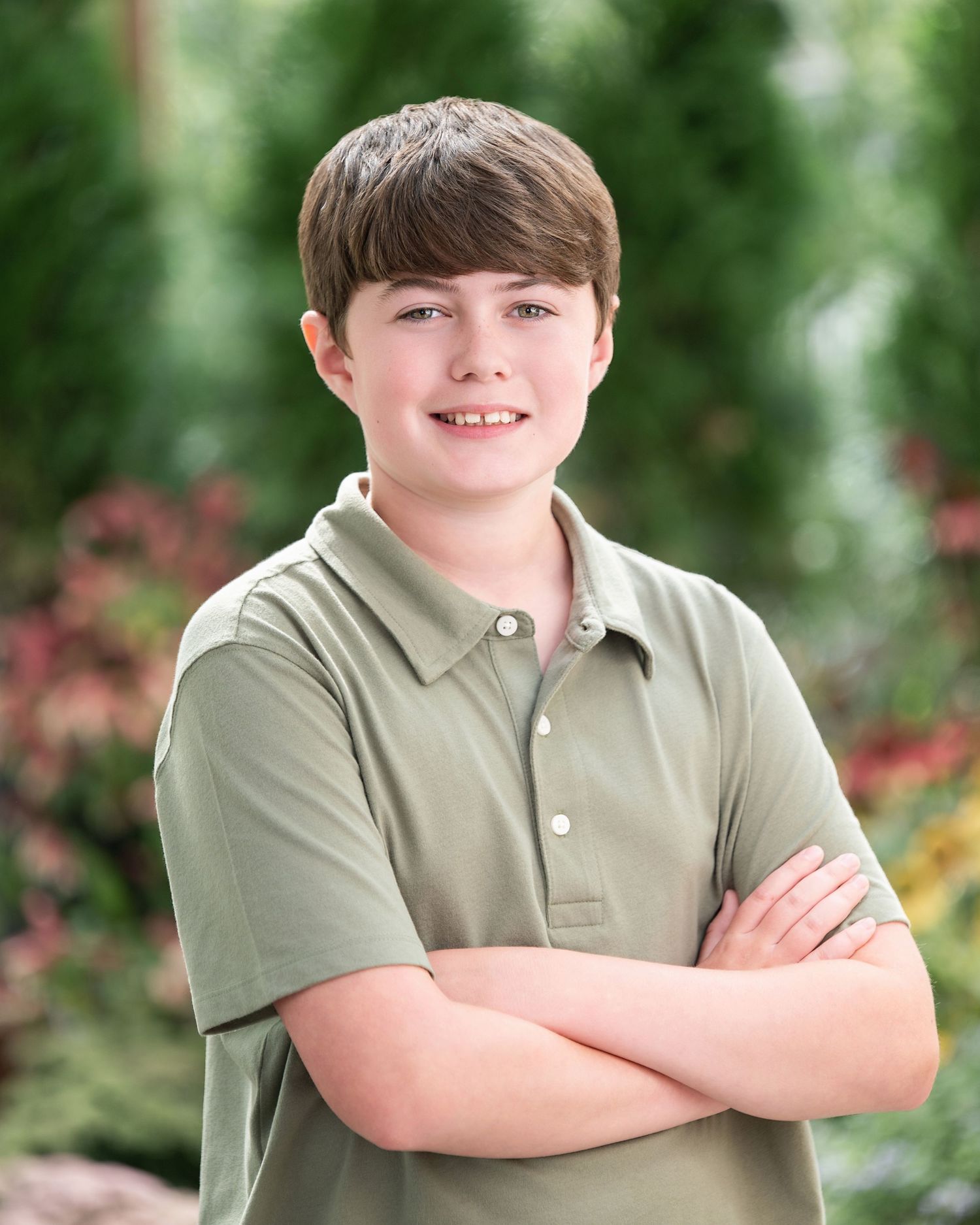 Smiling young boy with brown hair wearing a sage green polo shirt, arms crossed, posing outdoors with greenery background.