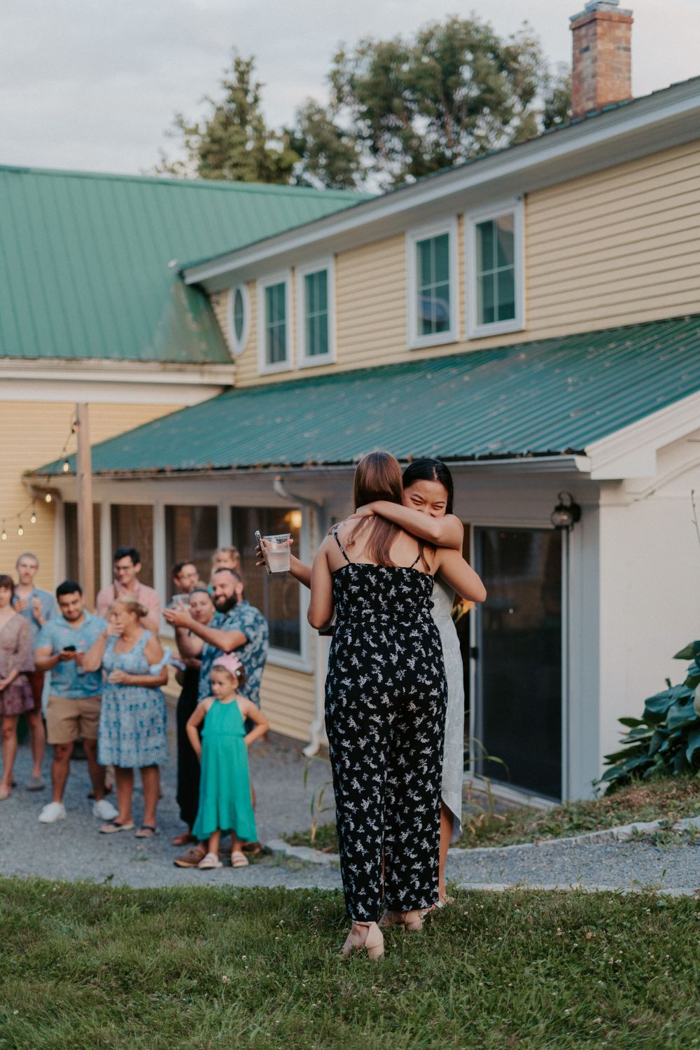 Two women embrace on a lawn while wedding guests applaud outside a yellow farmhouse at dusk.
