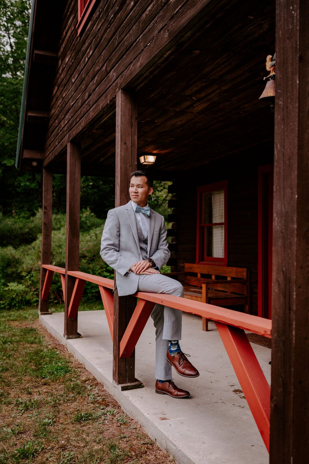 Dapper groom in grey suit and bow tie sitting on red porch railing of rustic wooden barn.