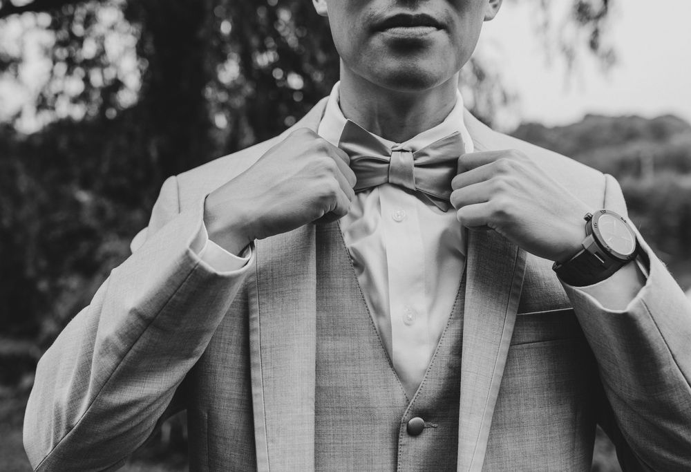 Man in light gray suit adjusting bow tie outdoors, wearing a watch, black and white wedding photo.