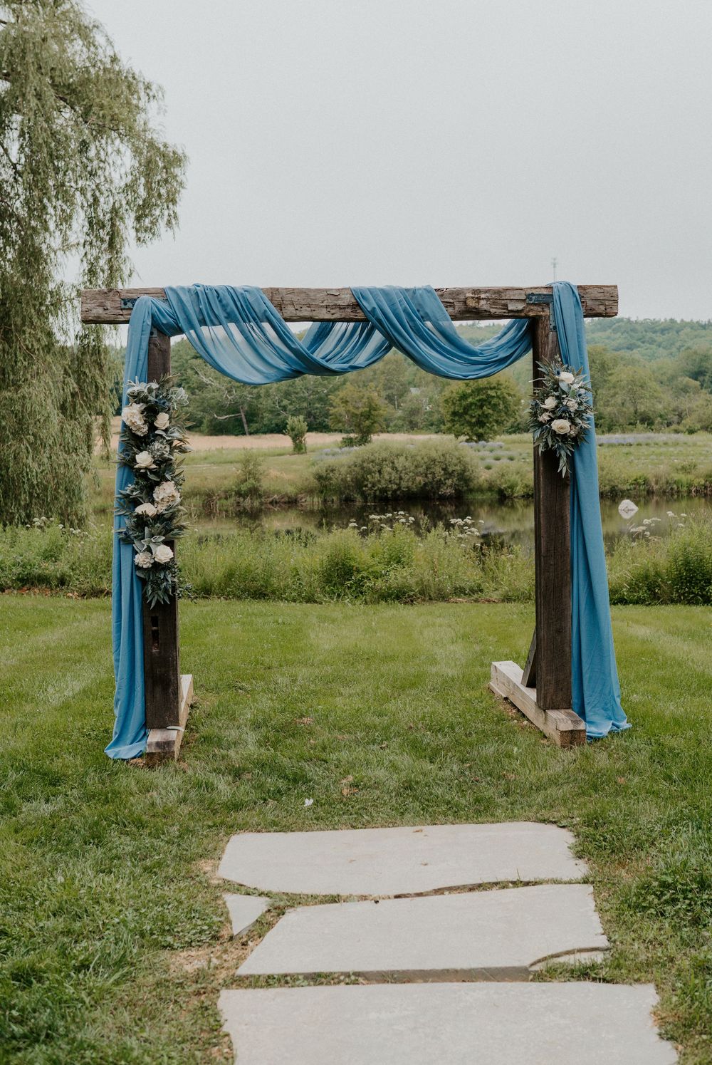 Rustic wooden wedding arch draped with blue fabric and white floral arrangements on a green lawn.