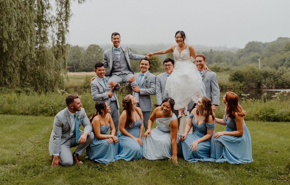 Wedding party group photo with bride being lifted by groomsmen on lush green lawn.