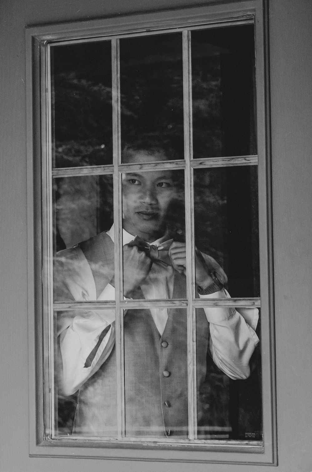 Black and white photo of a well-dressed man in a vest adjusting his bow tie while standing behind a multi-pane window.