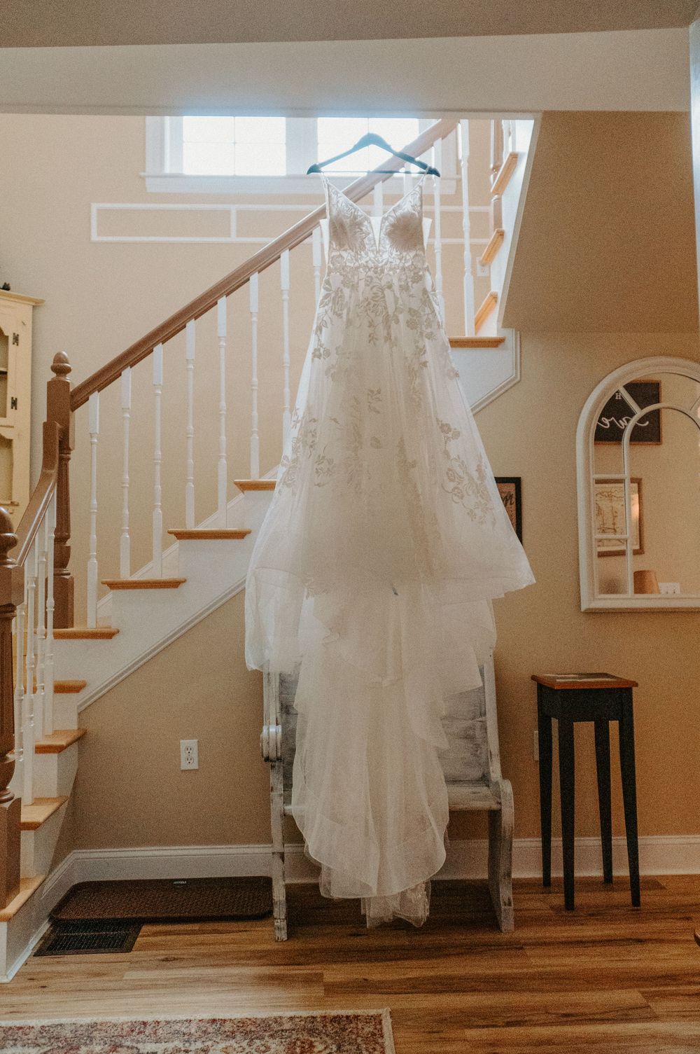 Elegant white lace wedding gown hanging on a staircase banister in a warmly lit home foyer.