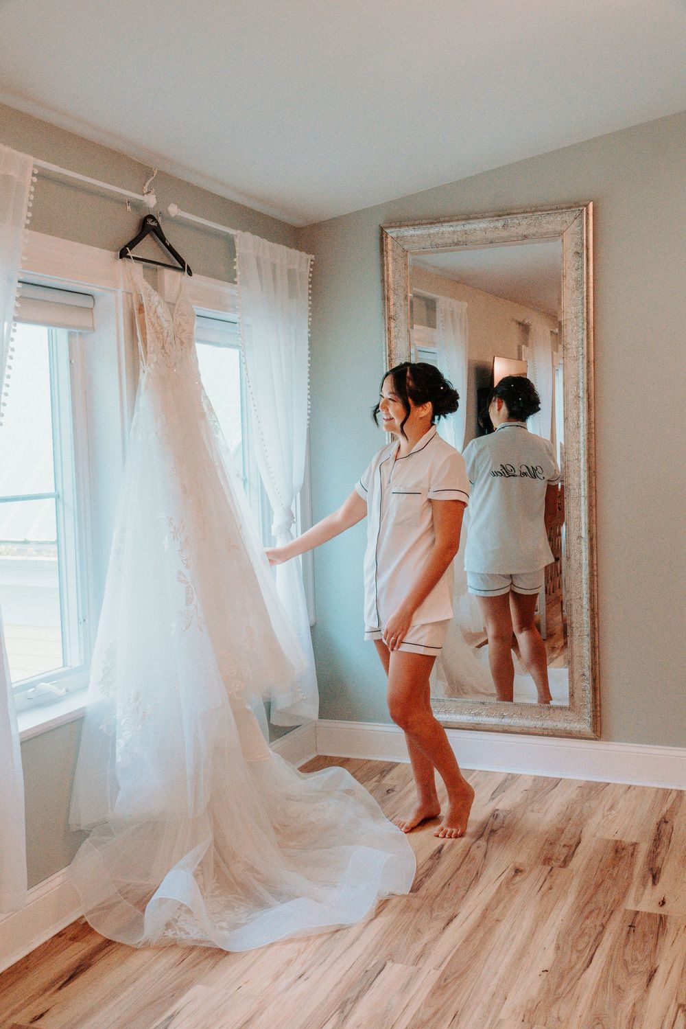 Bride in pajamas admiring her wedding gown hanging by window reflected in large floor mirror.