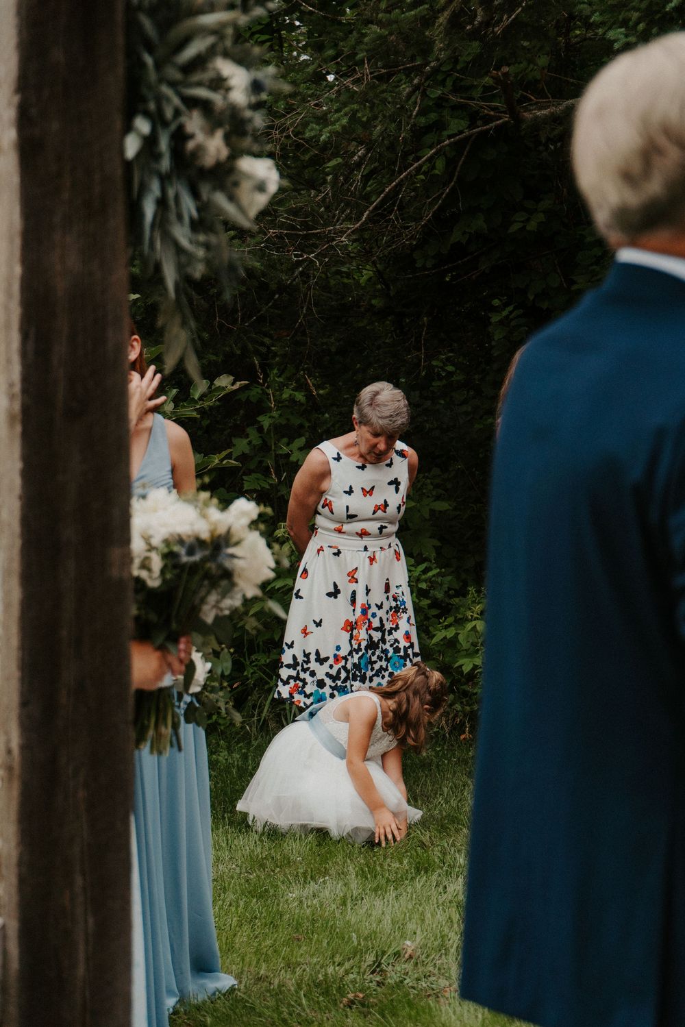 Flower girl bending down during outdoor wedding ceremony with bridesmaids in blue dresses and guest in butterfly print dress.