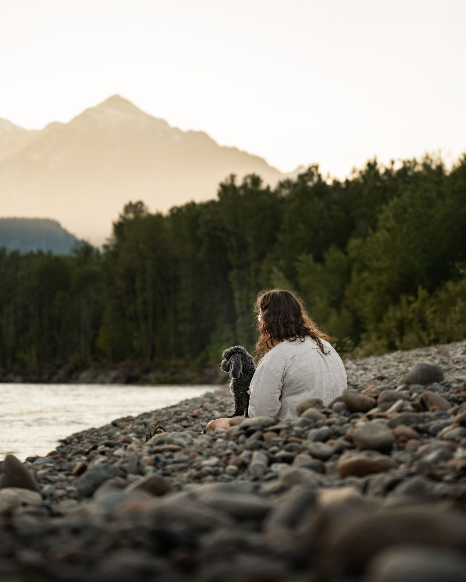 Woman and black dog sitting on rocky riverbank at golden hour with mountains and forest in background.
