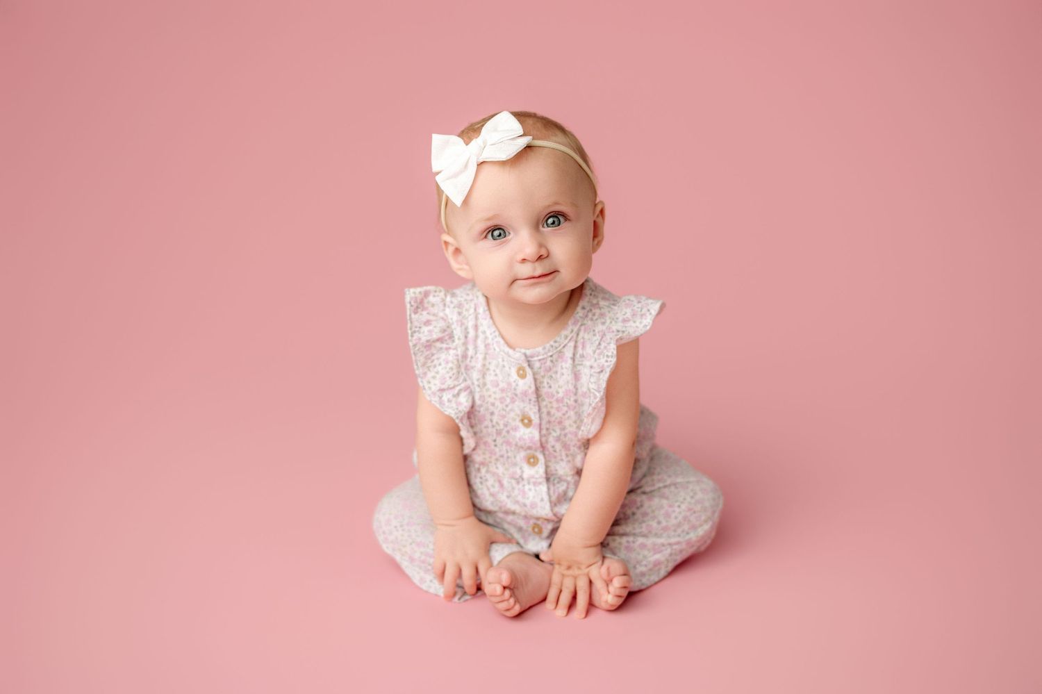 Adorable baby girl in white romper and bow headband sitting on pink background for professional portrait.
