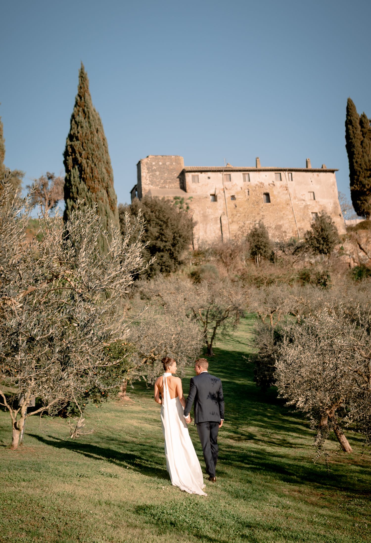 Bride and groom walk through olive grove toward historic stone Tuscan castle under clear blue sky.
