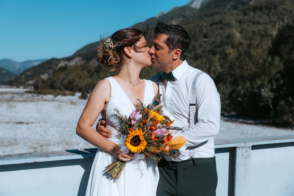 Bride and groom sharing a kiss outdoors in winter, bride holding sunflower bouquet with snowy mountains behind.