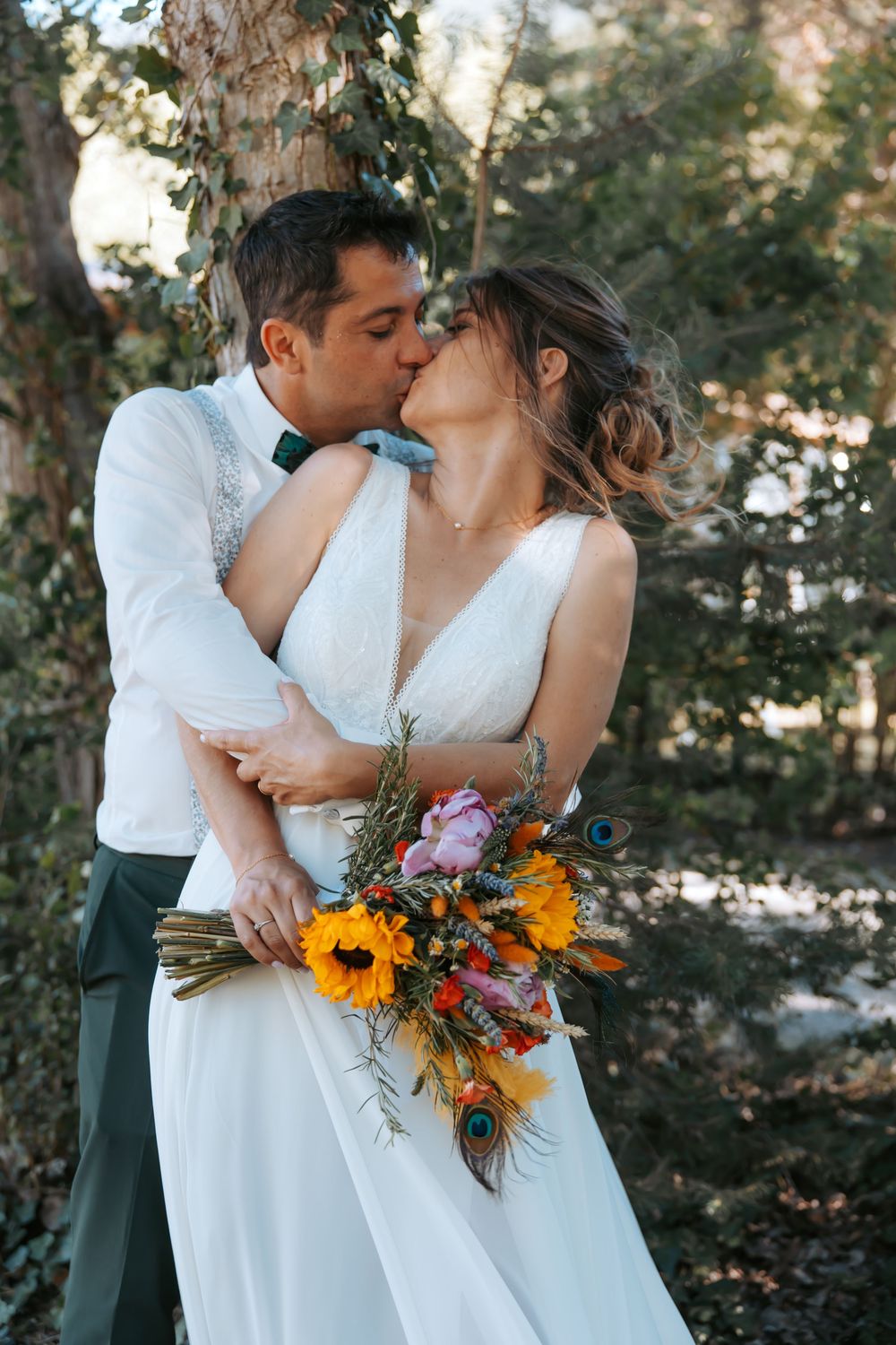 Bride and groom share a kiss outdoors, she holds a vibrant bouquet of sunflowers and peacock feathers in a forest setting.