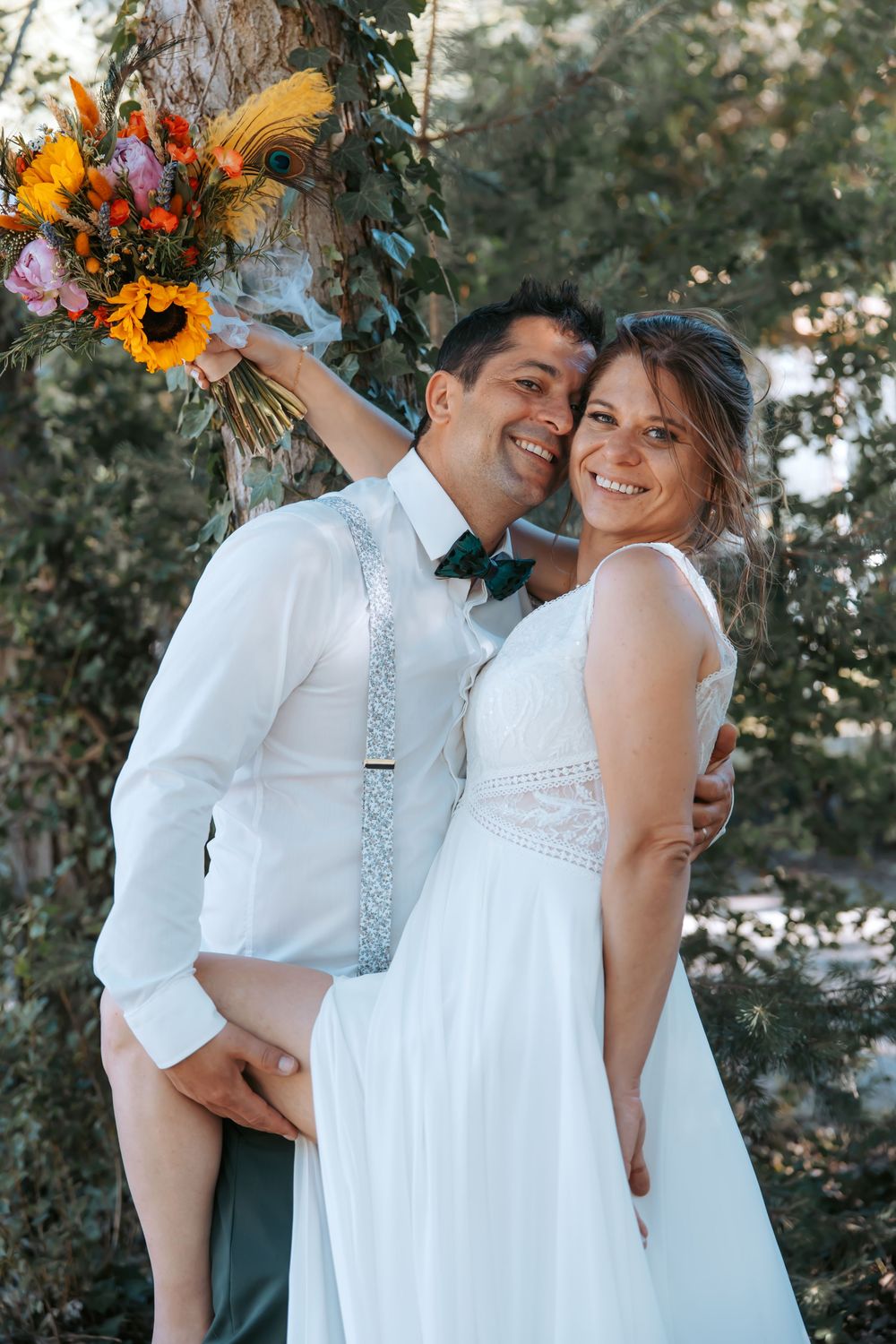 Happy newlyweds smiling outdoors, bride in white gown holding colorful sunflower bouquet, groom in white shirt with teal bow tie.