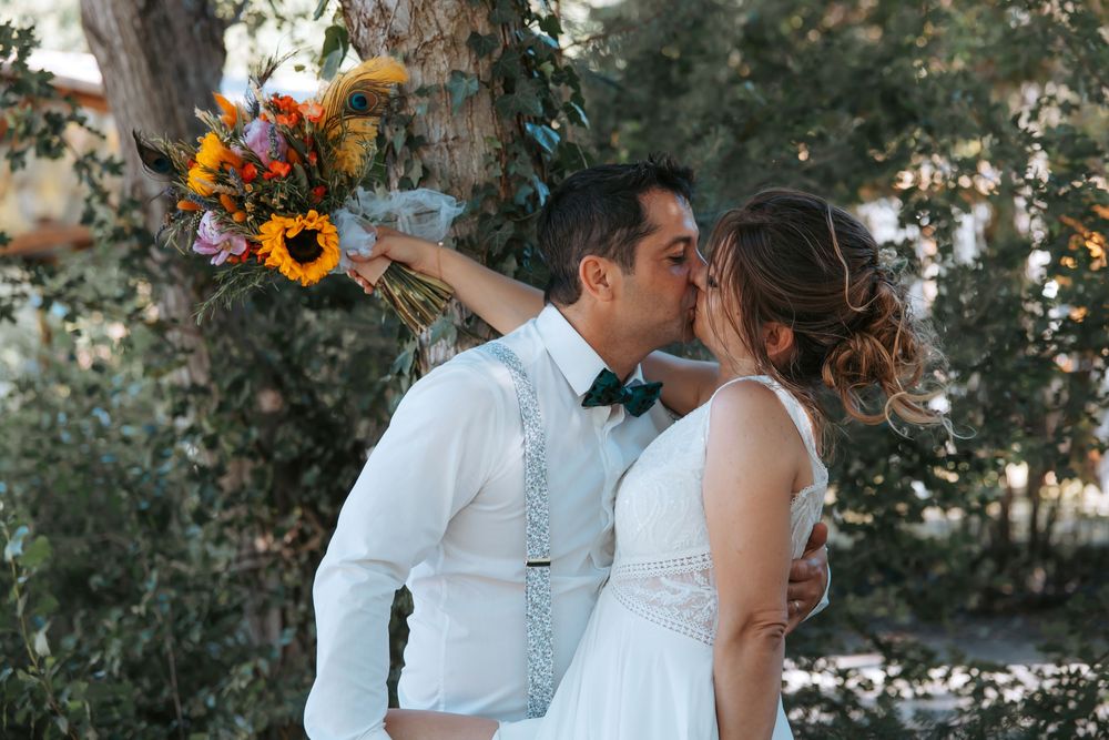 Newlywed couple sharing a kiss outdoors, bride holding vibrant sunflower bouquet, groom in white shirt with bow tie.