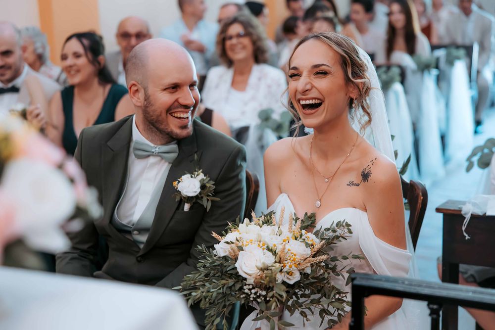 Joyful bride and groom laughing together at wedding ceremony, bride holding white floral bouquet with greenery.