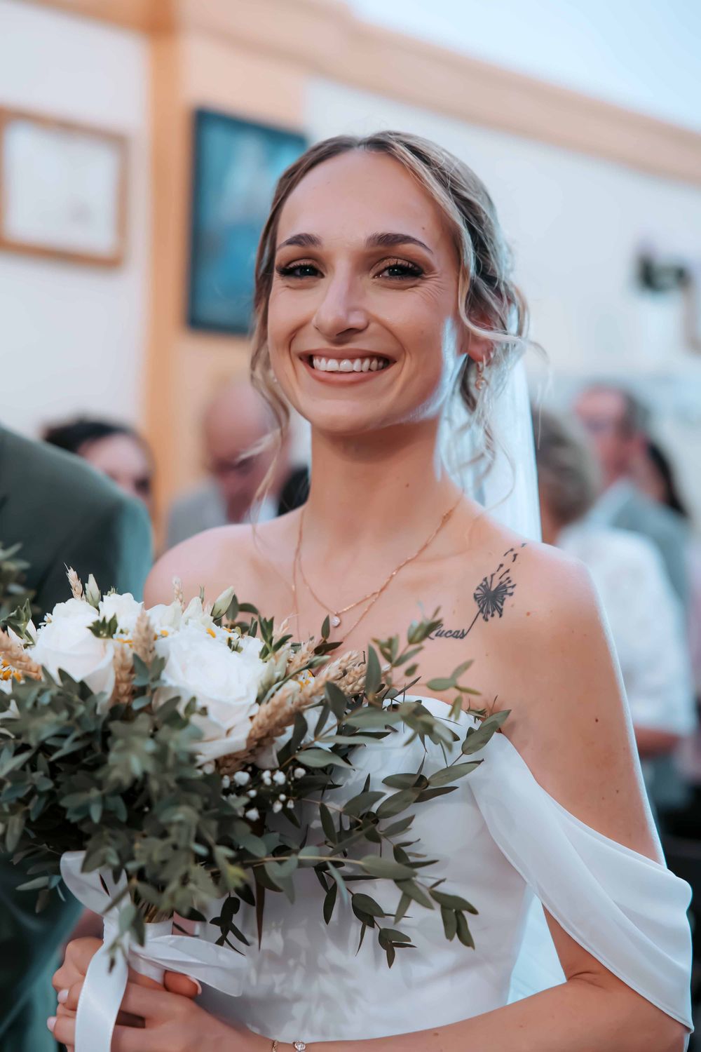Smiling bride in off-shoulder white dress holding white and green bouquet with tattoo visible on chest.