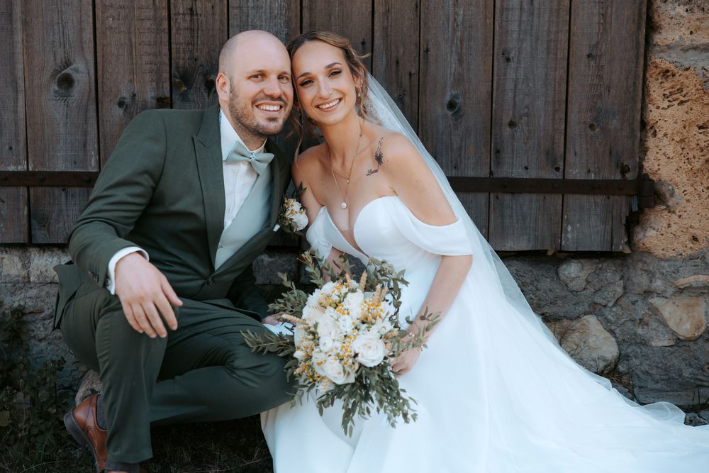 Happy bride and groom posing together in front of rustic barn, bride holding white floral bouquet.