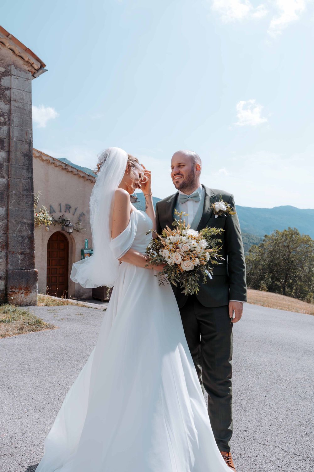 Bride and groom walking outdoors near a stone chapel with mountain views on their wedding day.