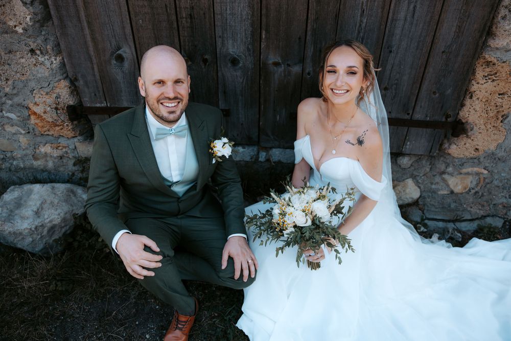 Bride and groom sitting together against rustic stone wall, bride holding white and yellow bouquet in elegant off-shoulder wedding gown.