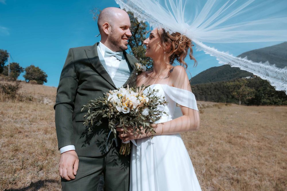 Bride and groom smiling at each other outdoors on their wedding day, with a flowing veil and rustic mountain backdrop.
