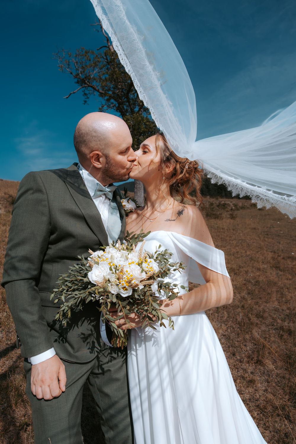 Bride and groom kissing outdoors with veil blowing in wind, holding white floral bouquet against blue sky.