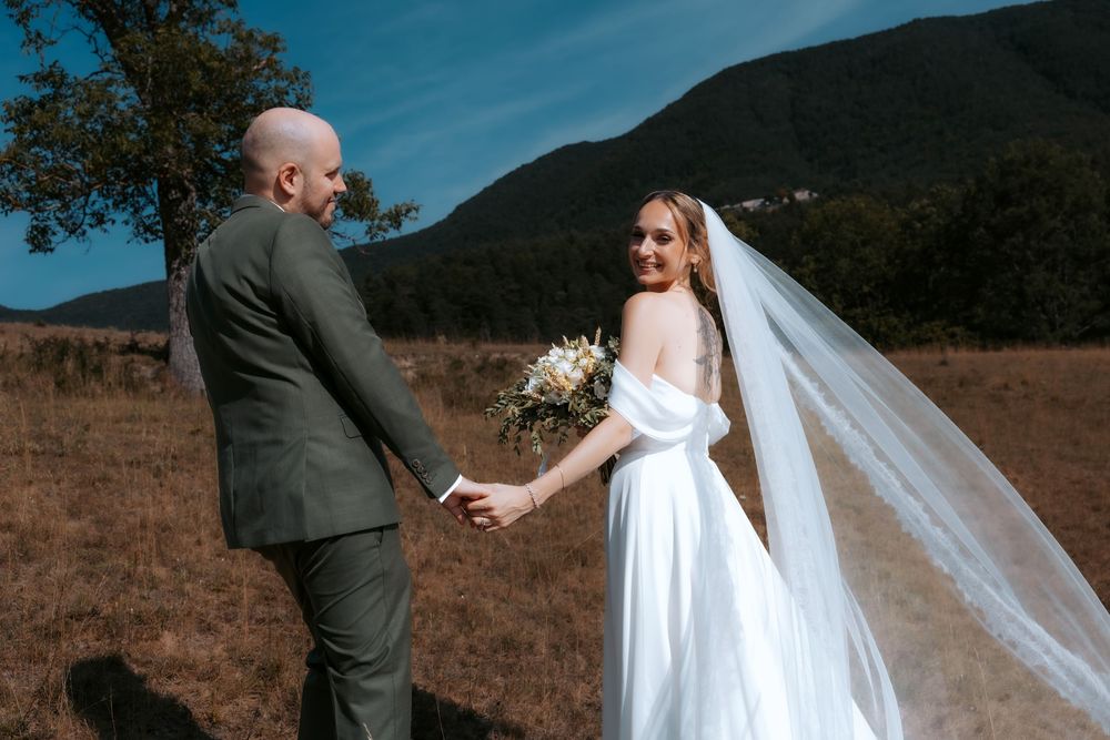 Bride in white gown and veil holds bouquet while groom in gray suit holds her hand in mountain meadow.