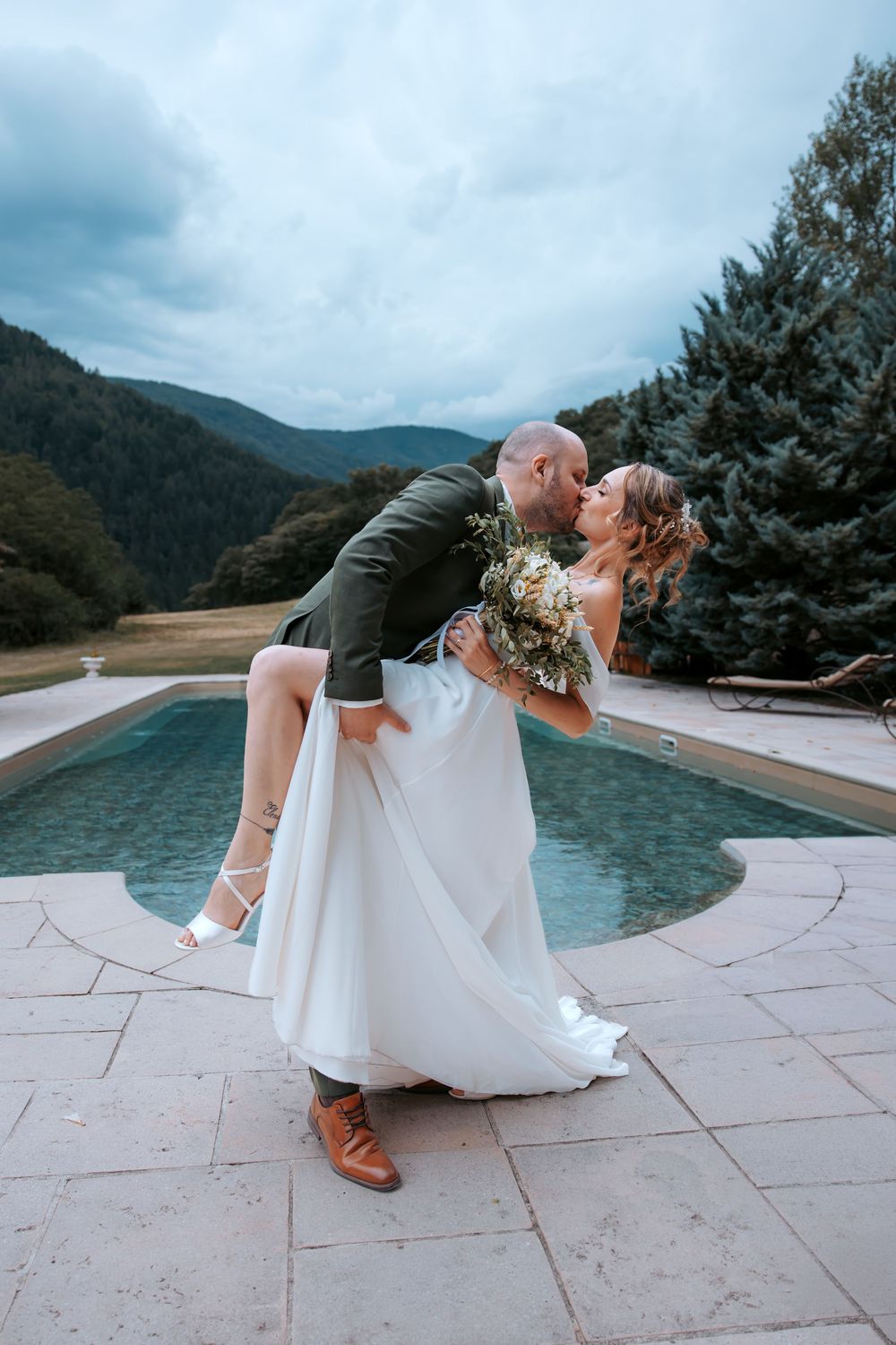 Groom dips bride for a romantic kiss by a pool with mountain scenery on their wedding day.