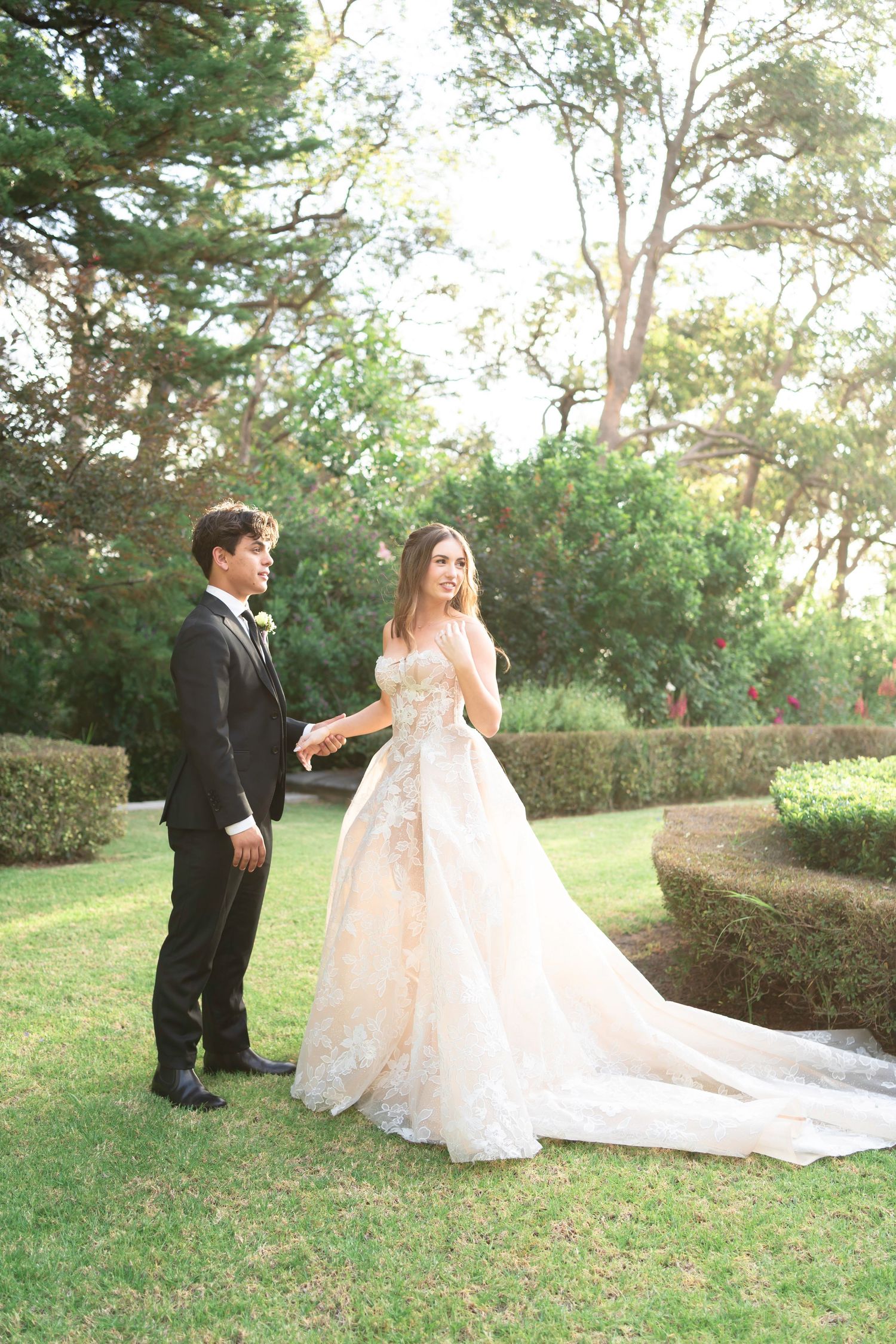 Wedding couple poses playfully in a sunlit garden setting with flowing dress train.