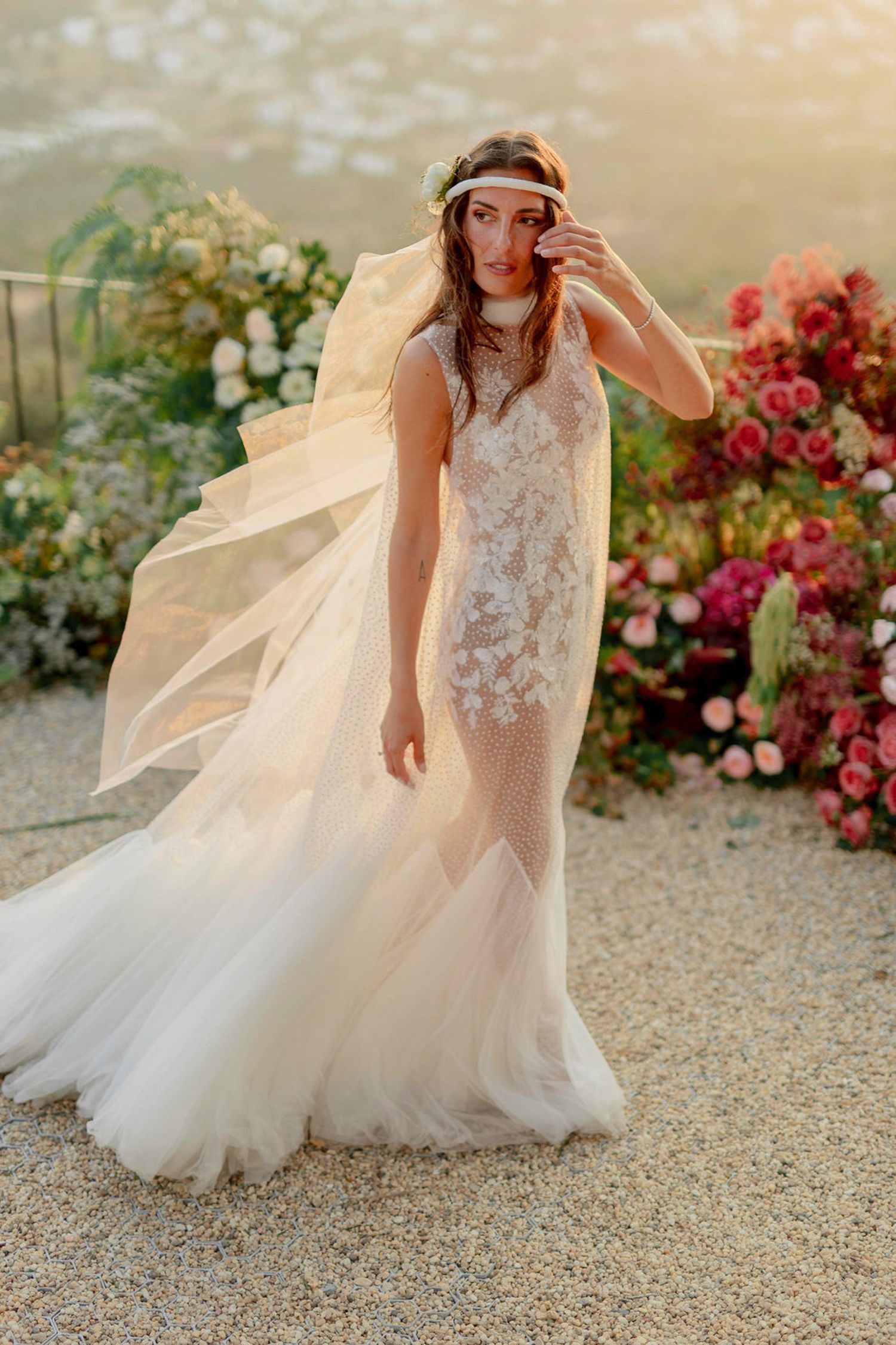 Bride in sheer lace mermaid wedding gown with floral headpiece, posing outdoors among colorful blooms. Sunset times in Bodrum, Turkiye