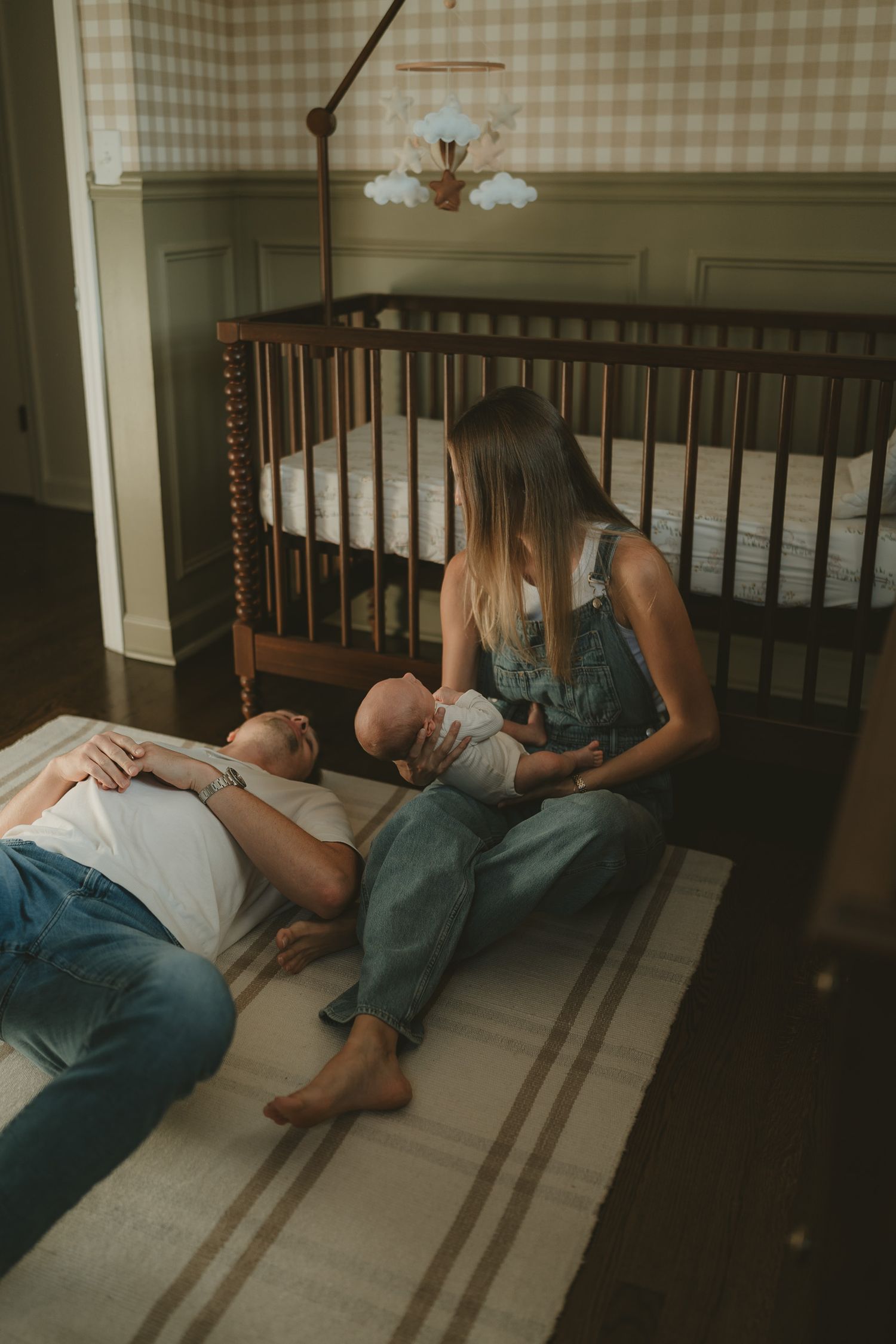 New parents sit on nursery floor with newborn baby beside a wooden crib with gingham wallpaper.