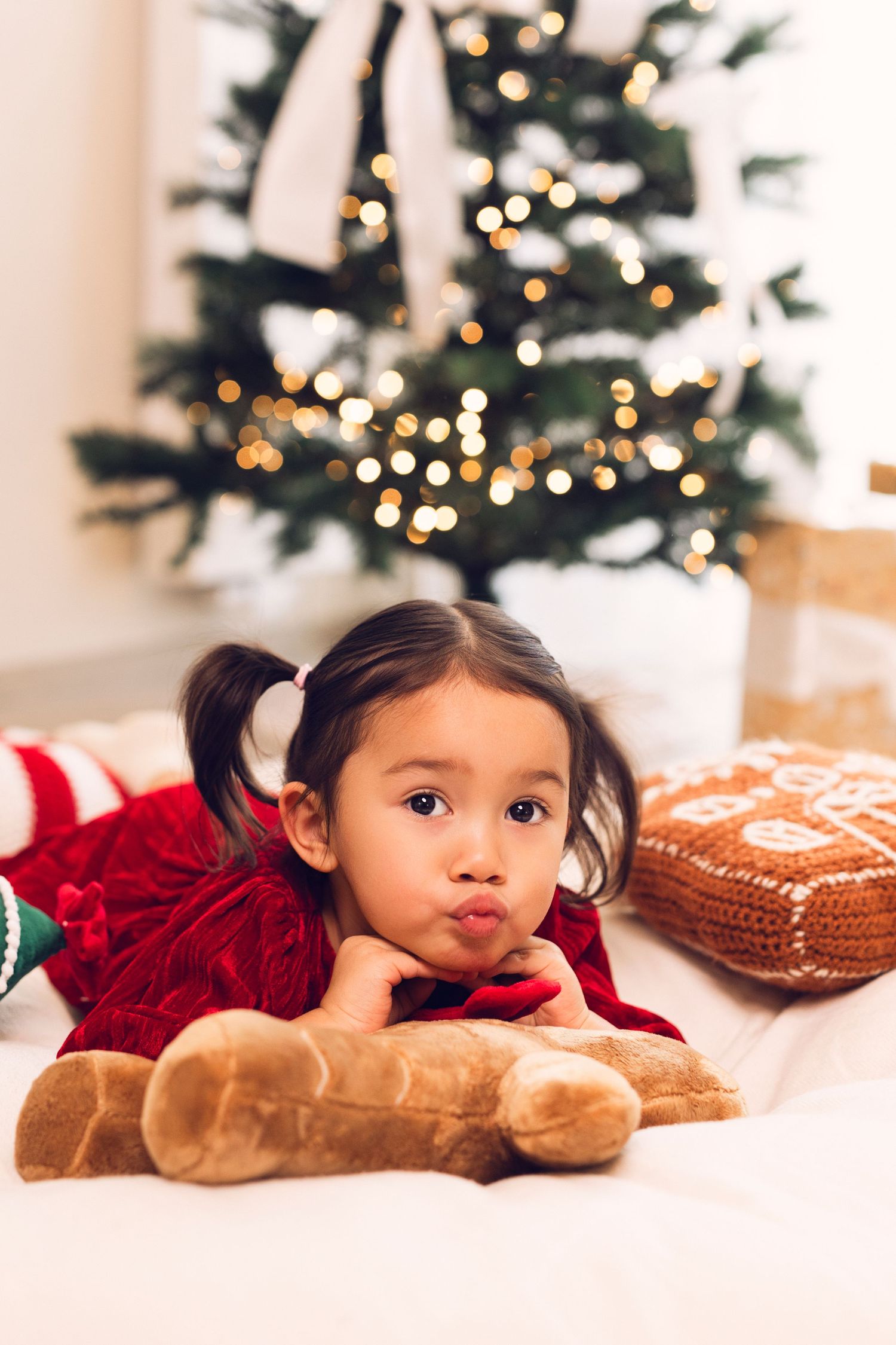 Young girl in red dress lying with teddy bear in front of decorated Christmas tree with golden lights.