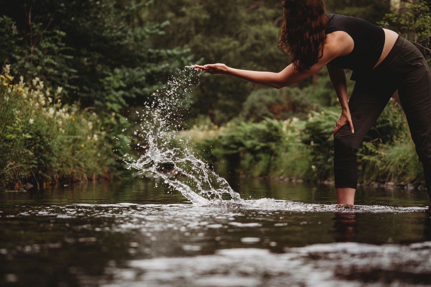 Woman splashing water in a serene forest river, wearing black outfit, surrounded by lush green trees.