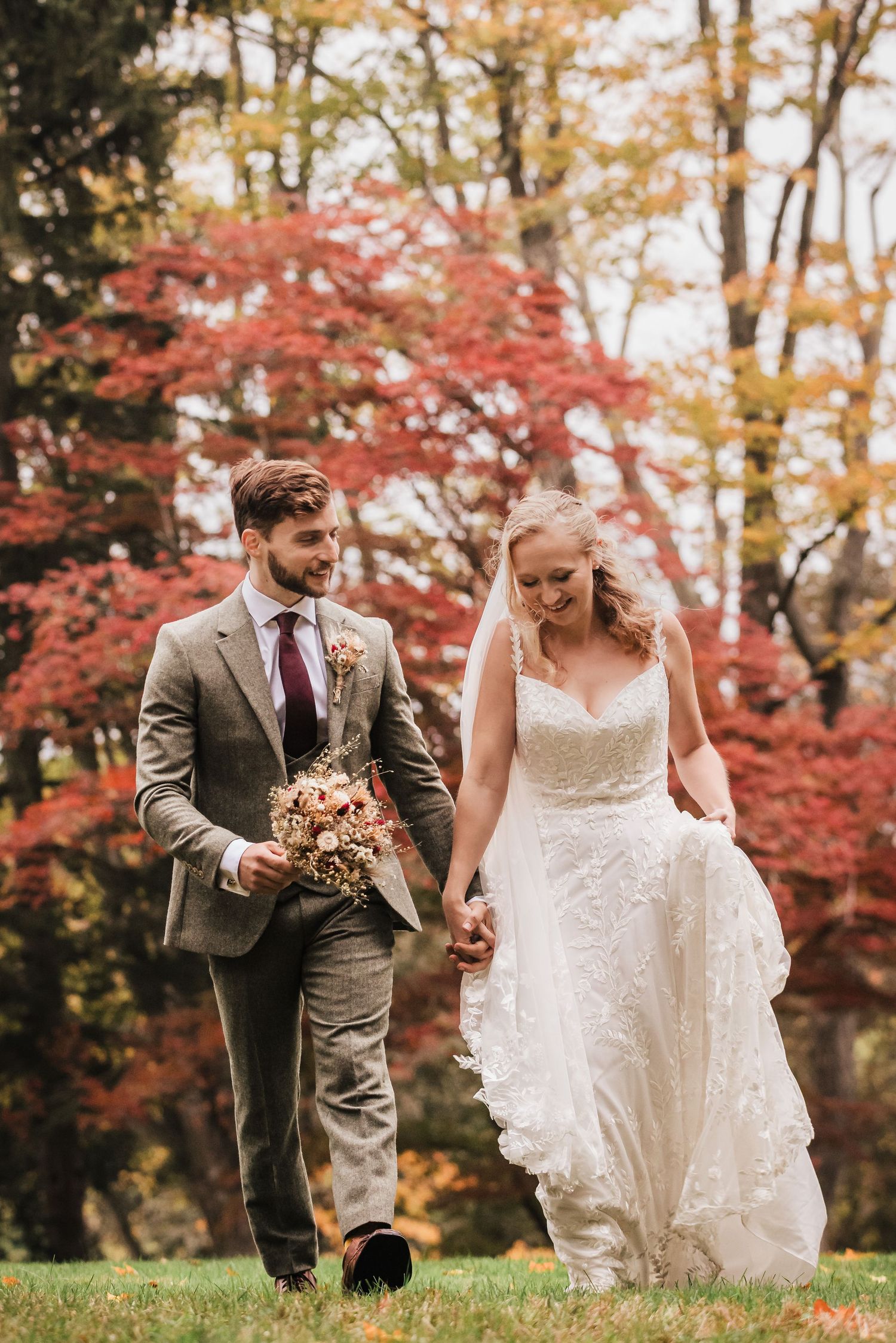 Bride and groom walking hand in hand through autumn grounds, holding bouquet. Gregory Hitchcock Photography