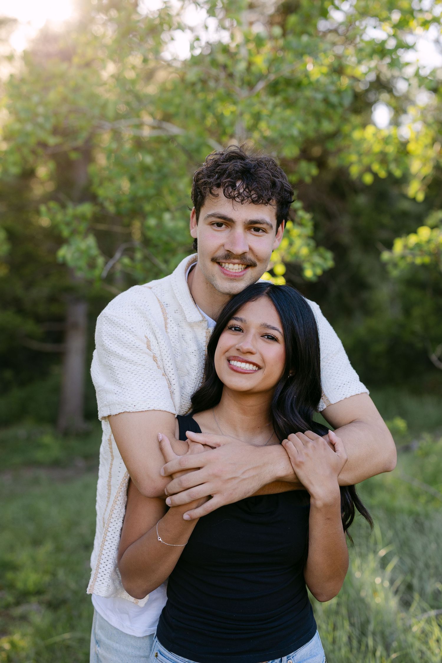 Couple embracing on a mountain trail surrounded by lush green forest with a rocky peak in the background.