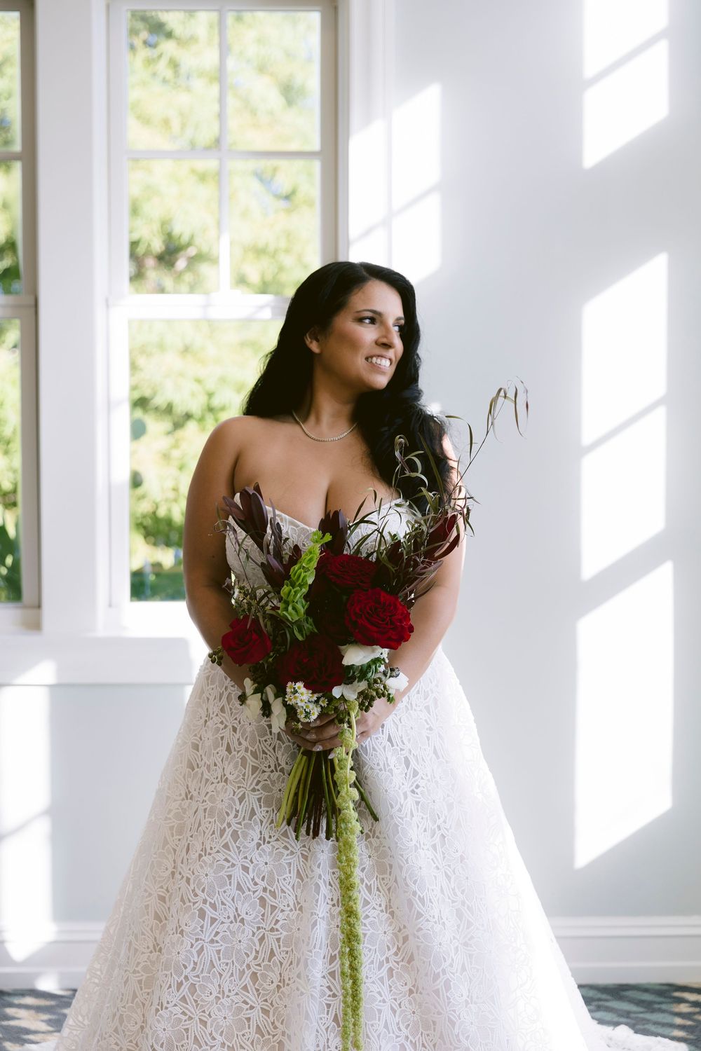 Bride in strapless lace ball gown holding red rose bouquet stands by sunlit window, smiling softly.