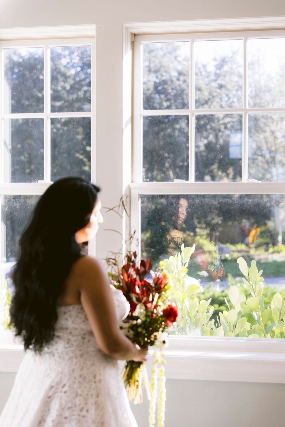 Bride in lace gown holding colorful bouquet gazing out sunlit window on wedding day.