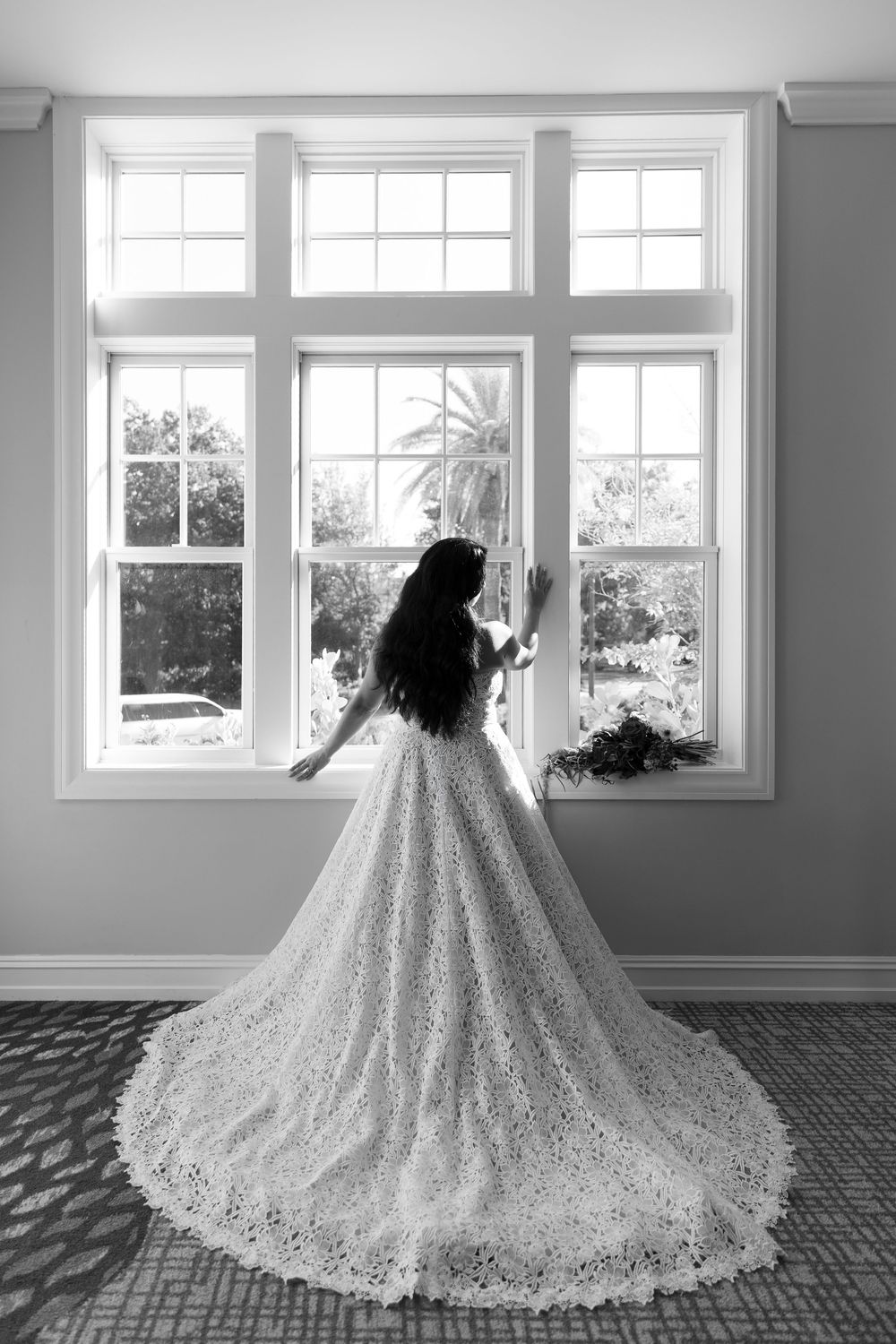 Bride in elegant lace ball gown with long train stands before large window in black and white wedding photo.