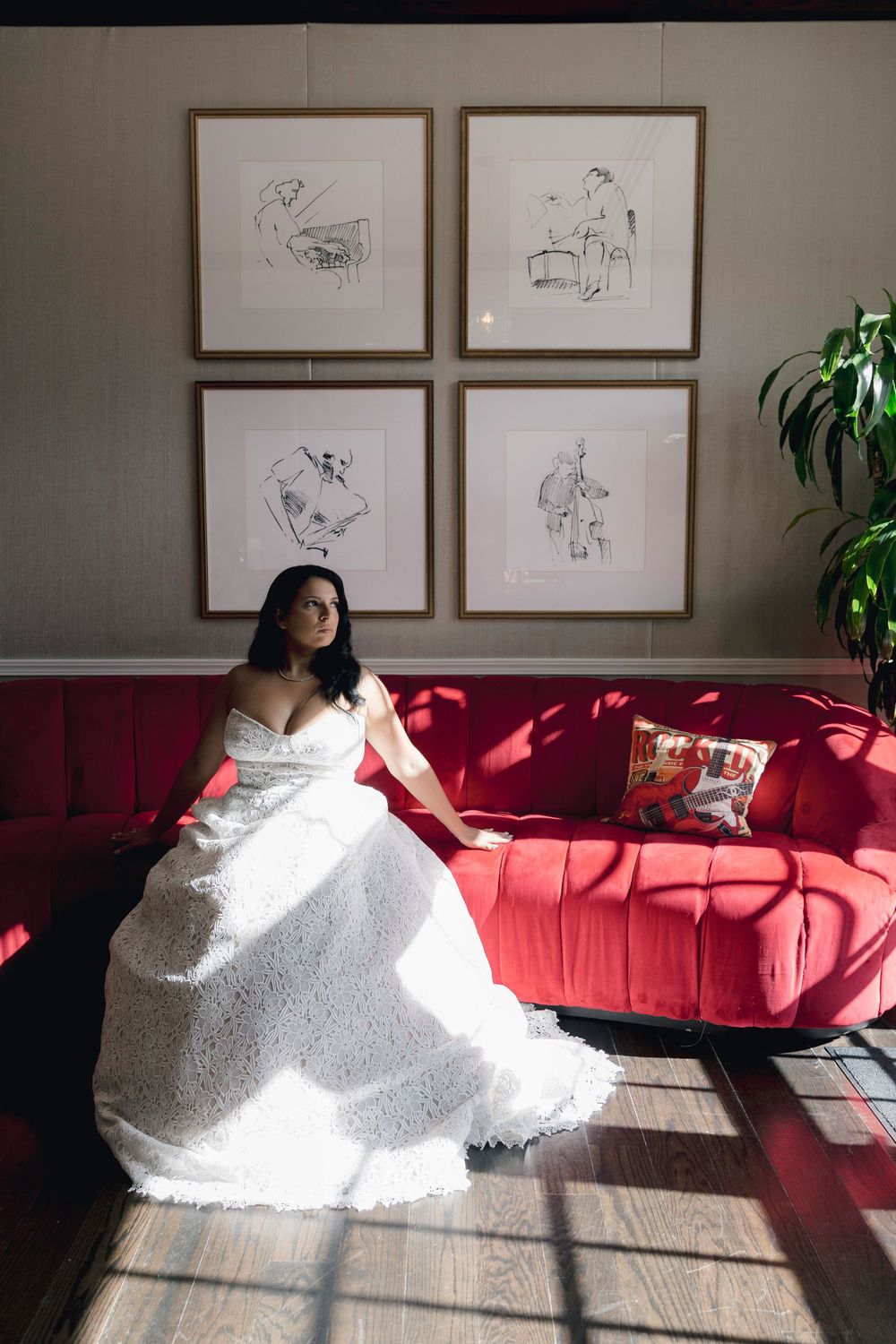 Bride in white lace gown seated on red velvet sofa with framed sketches on wall behind her.
