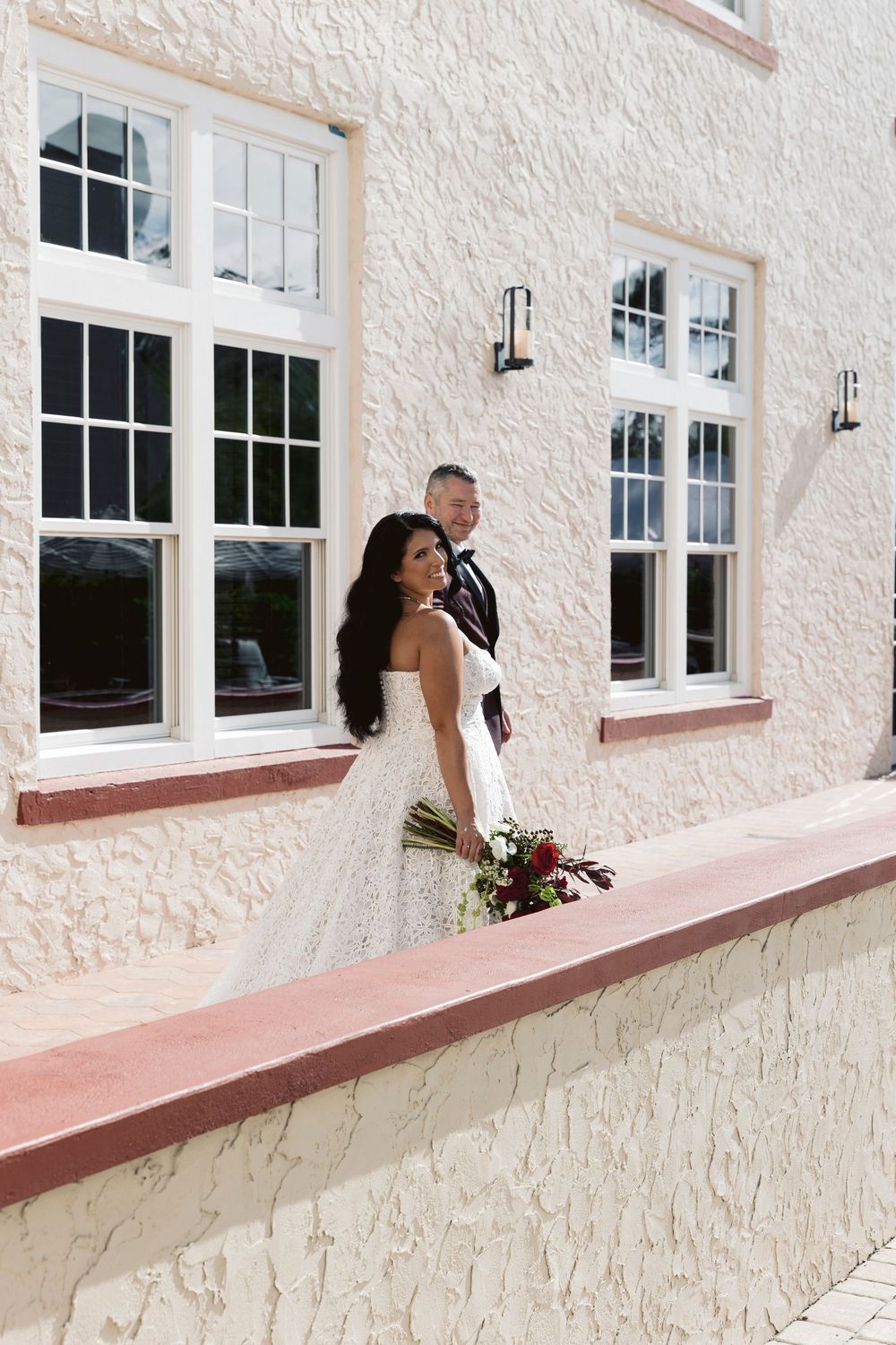 Bride in white lace gown holding bouquet stands before groom in suit outside elegant building with white windows.